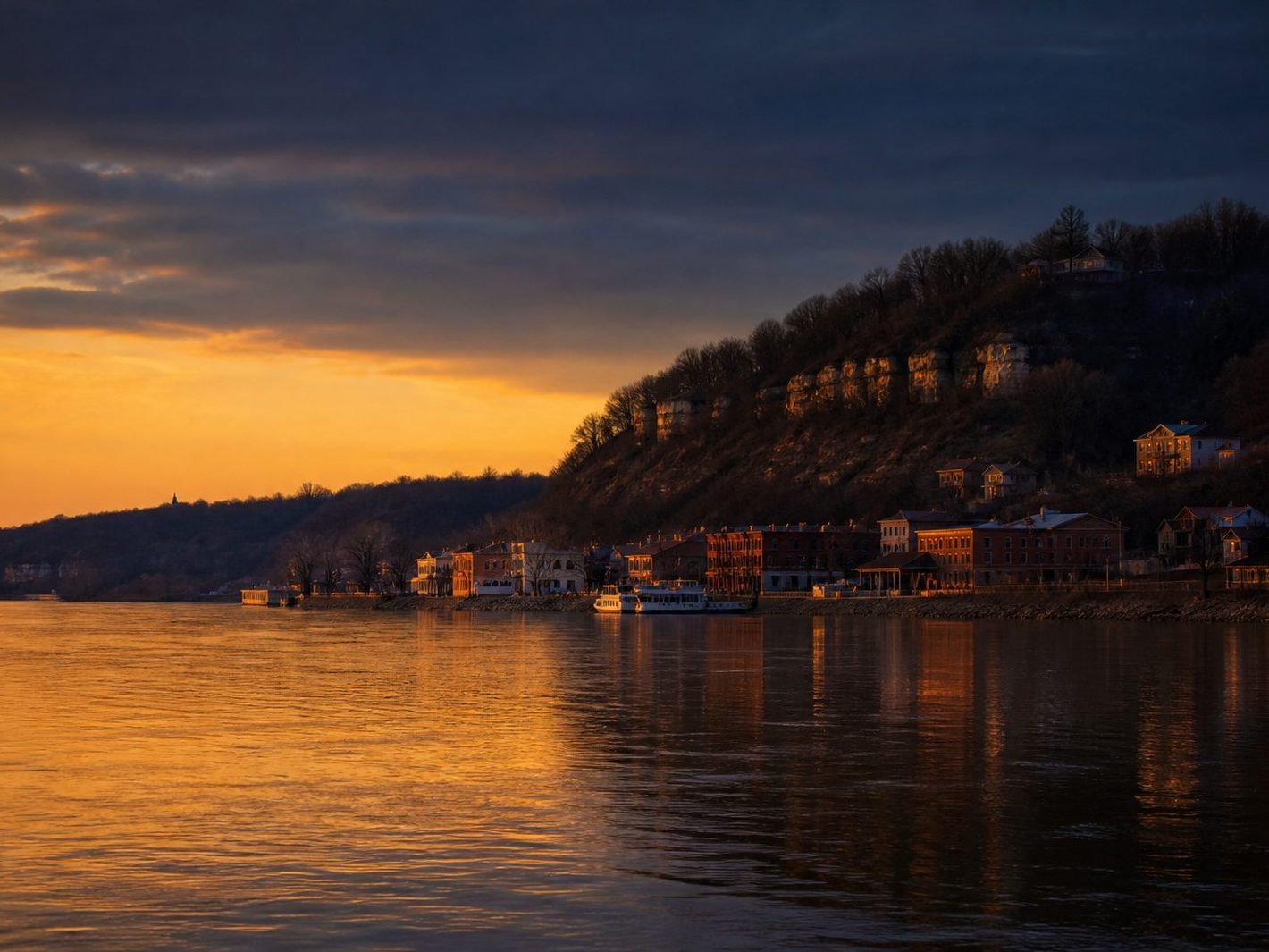 The Missouri River sliding past the St. Charles riverfront at dusk, brick storefronts and the bluff lit orange under a dark sky