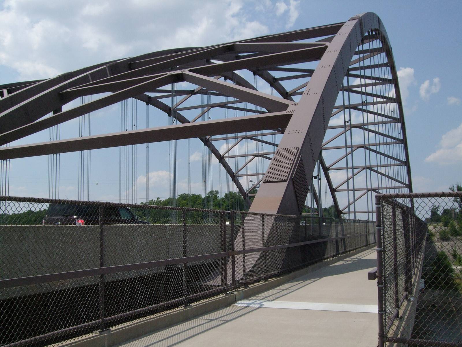 The steel tied-arch of the Route 364 bridge over the Missouri River, seen from the fenced trail lane beside the roadway on a summer afternoon